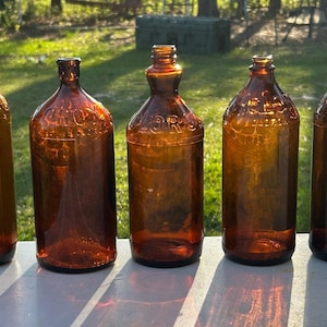 May include: Five antique amber glass bottles of varying shades and shapes are displayed outdoors. The bottles have embossed text and are arranged in a row. The bottles are likely vintage or collectible items, showcasing a range of designs and colors.