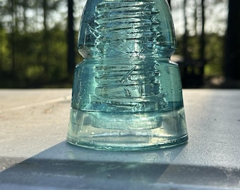 May include: A vintage, aqua-colored glass insulator with a rounded top and a tiered base. The glass has a textured surface and visible imperfections, suggesting age. The insulator is sitting on a white surface, with a blurred forest background.
