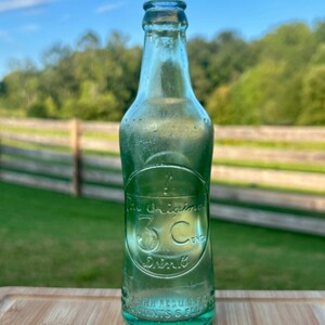 May include: A vintage light green glass soda bottle with an embossed circular logo featuring the text The Original 3 Cent Drink. The bottle has a narrow neck, a flared rim, and additional embossed text near the base reading Reg U.S. Pat Off.