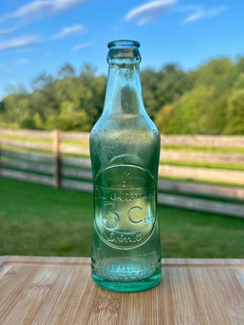 May include: A vintage light green glass soda bottle with an embossed circular logo featuring the text The Original 3 Cent Drink. The bottle has a narrow neck, a flared rim, and additional embossed text near the base reading Reg U.S. Pat Off.
