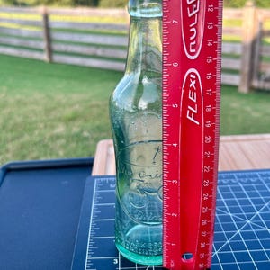 May include: A clear, light green glass bottle with a narrow neck and a rounded rim. The bottle features embossed text that reads 'The Original 30 Drink'. A red Flexi ruler is placed vertically next to the bottle, showing a height of 8 inches.