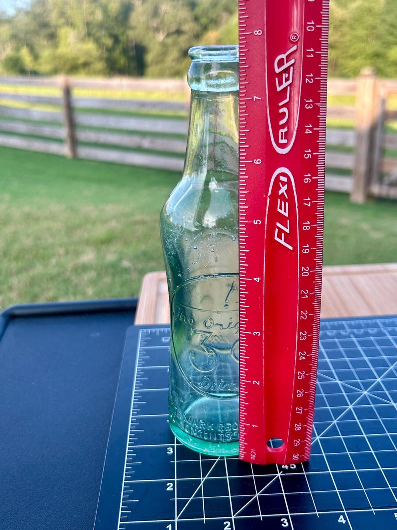May include: A clear, light green glass bottle with a narrow neck and a rounded rim. The bottle features embossed text that reads 'The Original 30 Drink'. A red Flexi ruler is placed vertically next to the bottle, showing a height of 8 inches.
