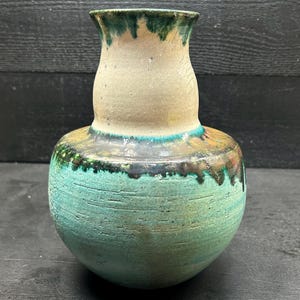 Op de afbeelding: Een handgemaakte keramische vaas met een bolle basis en een smalle hals. De vaas heeft een getextureerde turquoise body, een beige middengedeelte en een groen gerande rand. Een uniek decoratiestuk.