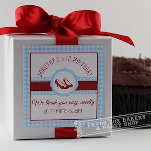 May include: A white cupcake box with a red ribbon and a blue and white gingham pattern label. The label says "Dorothy's 5th Birthday" and "We thank you very sweetly September 25, 2014". The box is sitting next to a chocolate cupcake with chocolate sprinkles.