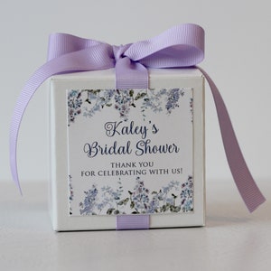 May include: A white gift box with a lavender ribbon tied in a bow. The box has a square label with a floral design and the text "Kaley's Bridal Shower Thank You for Celebrating with Us!"