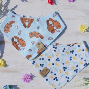 May include: Two baby bibs with different patterns. One is light blue with cartoon otters, shells, and hearts. The other is white with blueberries, lemon slices, and leaves. Both bibs have a tag that says "MAGNOLIA".