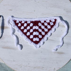 May include: A handmade crochet bandana in a triangular shape. The bandana features a burgundy and white checkered pattern with a scalloped white border. White ties extend from the corners for securing. The bandana is displayed on a white wooden surface.
