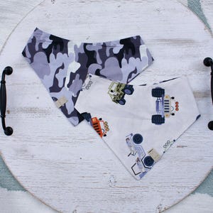May include: Two triangular pet bandanas. One is gray camouflage, the other is white with colorful illustrations of trucks and jeeps. The bandanas are displayed on a white wooden tray with black handles.