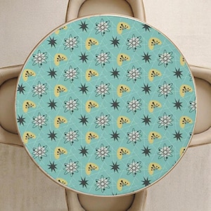 May include: A round table featuring a light blue surface with a repeating pattern of yellow boomerangs, dark gray starbursts, and white atomic symbols with thin gray lines connecting the elements, surrounded by four tufted beige chairs.