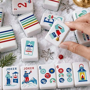 May include: A set of white rectangular mahjong tiles featuring colourful winter-themed illustrations, including skiers, lift tickets, mittens, scarves, and striped patterns. The tiles are arranged on a white surface with pine branches and ornaments.