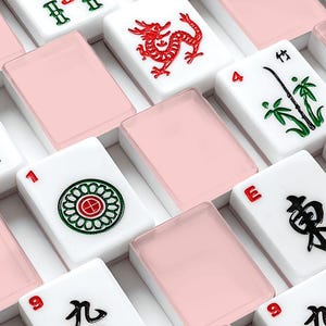 May include: A close-up shot of a Mahjong set with white tiles featuring red, green, and black symbols and numbers. Pink translucent tiles are interspersed between the white tiles. The set is arranged in a grid pattern.