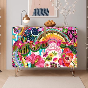 May include: A rectangular cabinet featuring a vibrant, multi-colored surface pattern with stylized floral motifs, stars, a crescent moon, and geometric shapes. The cabinet is supported by four thin, gold-toned metal hairpin legs and has two doors.