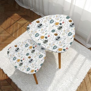 May include: Two white nesting tables with a whimsical forest pattern in shades of blue, green, yellow, and orange. The tables have a unique, organic shape and wooden legs.