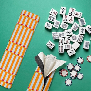 Puede incluir: Un juego de Mahjong completo con fichas blancas con símbolos negros y rojos, dispuestos sobre una superficie verde. Una funda a rayas naranja y blanca está abierta, revelando el interior. También se ven fichas de póquer.
