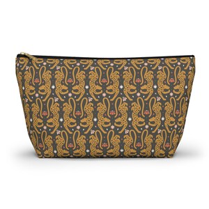 Peut inclure: Une pochette zippée avec un motif répété de guépards stylisés en or et noir. Le motif comprend des fleurs roses et des éclats d'étoiles blancs sur fond gris foncé. La pochette a une fermeture éclair noire.