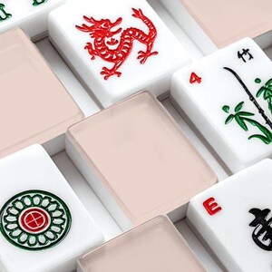 May include: Close-up of a set of Mahjong tiles. The tiles are white with various symbols and characters in red and green. Some tiles have a dragon design, while others feature bamboo or circular patterns. The tiles are arranged in a box with a light pink cover.