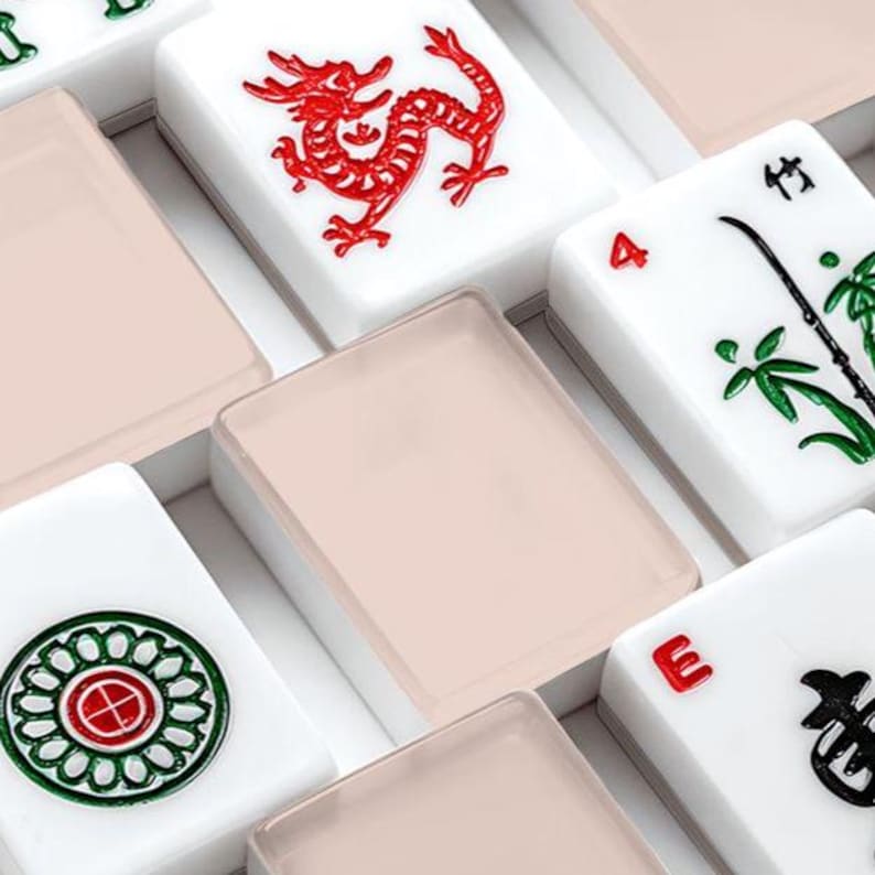 May include: Close-up of a set of Mahjong tiles. The tiles are white with various symbols and characters in red and green. Some tiles have a dragon design, while others feature bamboo or circular patterns. The tiles are arranged in a box with a light pink cover.