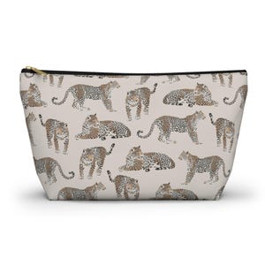 Peut inclure: Une pochette beige zippée avec un motif répété de léopards dans diverses poses. Les léopards ont des taches marron et sont placés sur un fond neutre. La pochette a une fermeture éclair dorée.