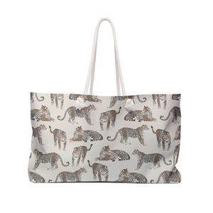 Peut inclure: Grand sac fourre-tout avec un motif répété de léopards bruns et beiges sur fond gris clair. Il est doté de poignées en corde blanche et de nœuds décoratifs blancs. Idéal pour le transport.