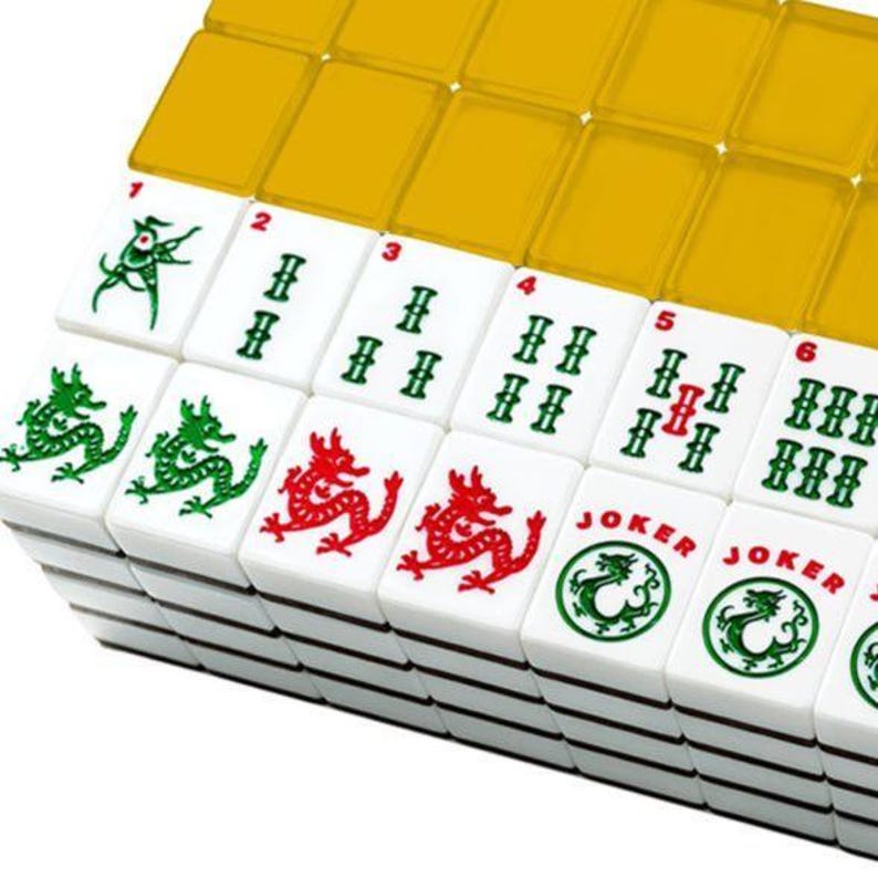 May include: A close-up of a Mahjong set with white tiles featuring green and red dragon designs, numbers, and the word "Joker". The tiles are stacked, with some covered by yellow translucent tiles.