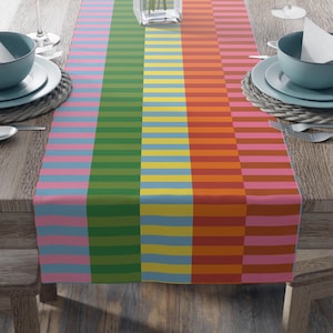 May include: A colorful table runner with a horizontal striped pattern in shades of pink, blue, green, yellow, orange, and brown. The runner is placed on a wooden table, with place settings visible on either side.