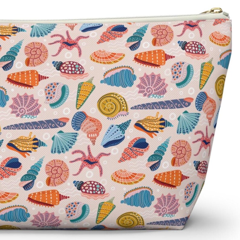 K&ouml;nnte beinhalten: Eine Kosmetiktasche mit einem sich wiederholenden Muster aus bunten Muscheln und Seesternen auf hellrosa Hintergrund. Die Tasche hat einen Rei&szlig;verschluss und einen cremefarbenen Rei&szlig;verschluss.