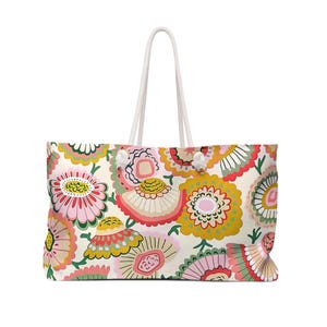 May include: A large tote bag featuring a repeating pattern of stylized flowers in shades of mustard yellow, soft pink, coral, and sage green on a cream background. The bag has two white rope handles attached with silver-toned metal grommets.