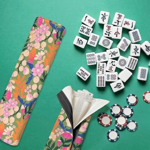 以下が含まれることがあります： 緑色の花柄の麻雀牌ケース。ファスナー付きで、内側が見えるように開いています。黒いシンボルと赤と青のチップが付いた白い麻雀牌が、緑色の表面に散らばっています。