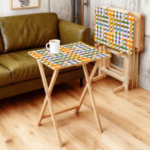 May include: A set of three folding TV trays with a colorful plaid pattern in orange, green, blue, pink, and yellow. The trays have natural wood legs and are set up in a room with a green sofa and a white mug on one tray.