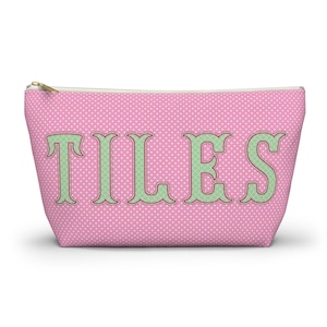 Puede incluir: Un estuche rosa con cremallera y un patrón de puntos blancos en toda la superficie. La palabra TILES está impresa en letras grandes de color verde claro con un borde verde oscuro y un sutil patrón de cuadrícula dentro de cada letra.