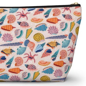 K&ouml;nnte beinhalten: Eine Kosmetiktasche mit Rei&szlig;verschluss und einem sich wiederholenden Muster aus bunten Muscheln, Seesternen und anderen Meereslebewesen auf hellrosa Hintergrund. Das Design umfasst orange, blaue, gelbe und violette Elemente. Der Rei&szlig;verschluss ist schwarz.