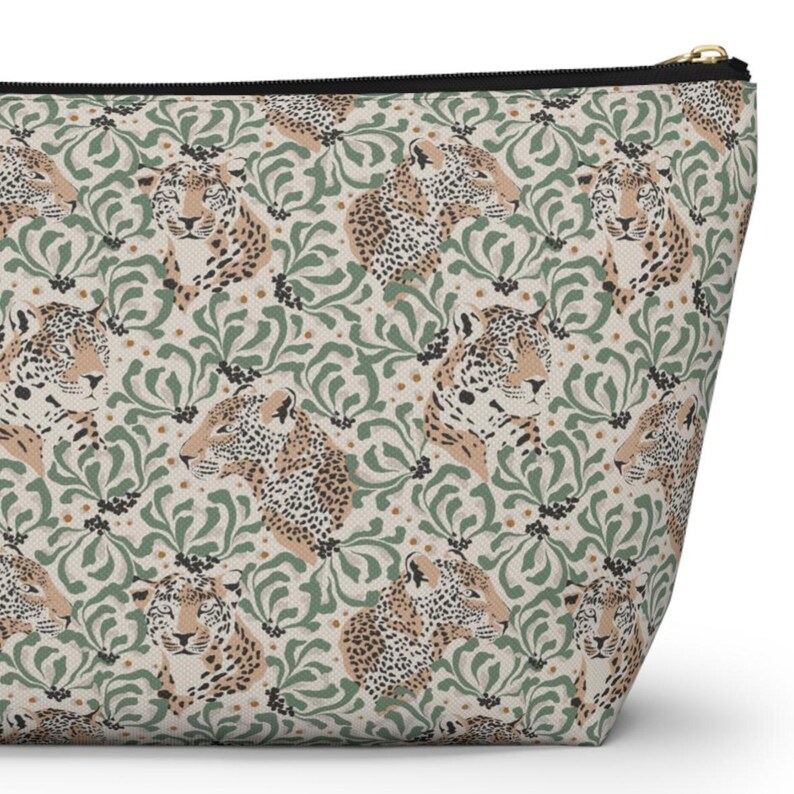 May include: A beige cosmetic bag with a repeating pattern of leopard faces and green floral designs. The bag has a black zipper and a gold-colored zipper pull. The overall design is a mix of animal print and botanical elements.