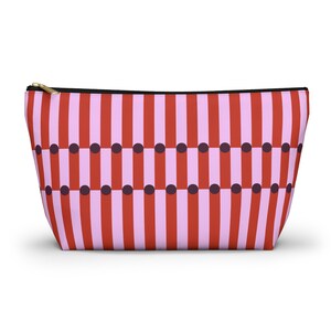 Puede incluir: Un neceser rectangular con un patrón repetitivo de rayas verticales rojas y rosa claro. Puntos morados oscuros están espaciados uniformemente entre las rayas. La bolsa tiene una cremallera negra y un tirador dorado.