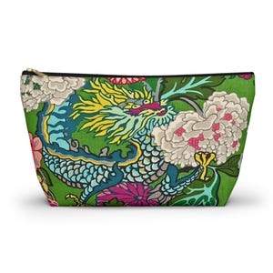 Könnte beinhalten: Eine grüne Reißverschlusstasche mit einem farbenfrohen Drachendesign mit gelben, blauen und türkisfarbenen Akzenten. Der Drache ist vor einem Hintergrund aus weißen und rosa Blumen und grünem Laub platziert. Die Tasche hat einen schwarzen Reißverschluss und einen goldfarbenen Zipper.