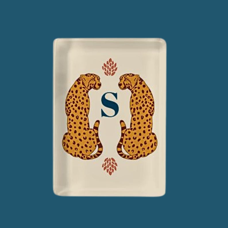 May include: A rectangular cream-colored tray featuring two symmetrical cheetah illustrations in gold and brown. Between the cheetahs is a large, dark blue capital letter S. Above and below the letter S are small, stylized red floral motifs.