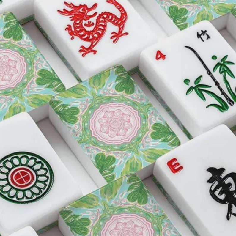 K&ouml;nnte beinhalten: Nahaufnahme eines Satzes wei&szlig;er Mahjong-Steine mit verschiedenen Symbolen und Zahlen in Rot, Gr&uuml;n und Schwarz. Einige Steine zeigen einen roten Drachen, Bambusst&auml;ngel und die Zahl 4. Das Set enth&auml;lt Steine mit einem floralen Design in Gr&uuml;n und Rosa.