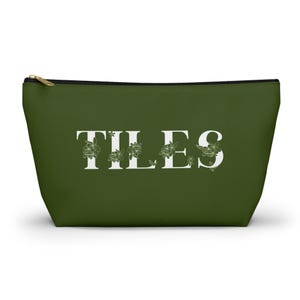 Peut inclure: Une pochette zippée vert olive avec le mot TILES en grandes lettres capitales blanches. Les lettres sont décorées d'un motif floral délicat aux lignes fines dans une teinte vert foncé, et la pochette possède une tirette de fermeture éclair dorée.