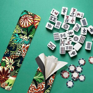 Peut inclure: Un long étui en tissu rectangulaire orné d'un motif vibrant de dragon et de fleurs dans des tons de vert, jaune, rouge et blanc, accompagné d'une collection de tuiles de mah-jong blanches avec des symboles noirs et rouges et des jetons.