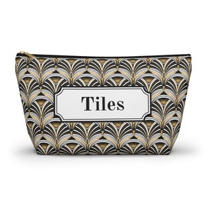 Puede incluir: Un estuche con cremallera que presenta un patrón repetitivo en forma de abanico en negro, blanco y dorado. Centrada en la parte frontal hay una etiqueta rectangular blanca con borde negro que contiene la palabra "Tiles" en tipografía serif.