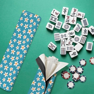 Puede incluir: Un estuche azul para fichas de Mahjong con un estampado de margaritas blancas, abierto para revelar una cremallera y el interior. Las fichas de Mahjong y las fichas de juego están esparcidas sobre una superficie verde.