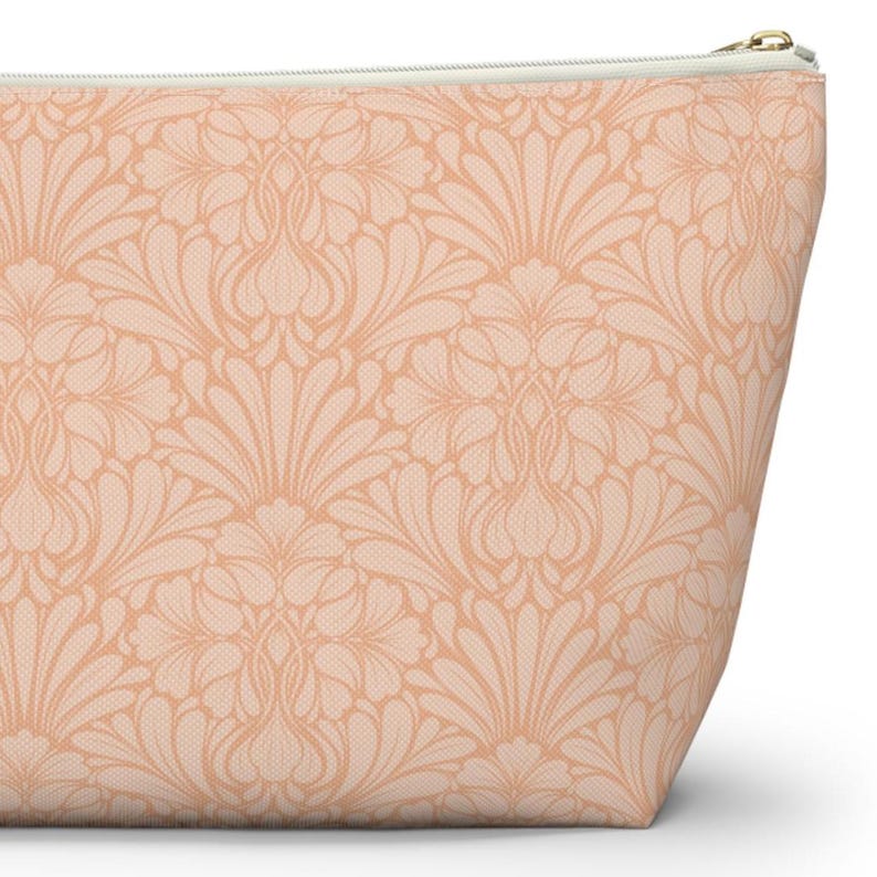 Op de afbeelding: Een perzikkleurige make-uptas met een bloemenpatroon. De tas heeft een herhalend ontwerp van gestileerde bloemen en bladeren in een lichtere perziktint. Een goudkleurige ritssluiting loopt langs de bovenrand.
