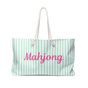 May include: A tote bag featuring a vertical mint green and white striped pattern. The word Mahjong is printed across the center in a bold, cursive pink font. The bag includes two white rope handles attached with knotted ends at the top.