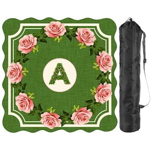 May include: A square picnic blanket with scalloped edges featuring a green textured background, a central cream circle with a floral letter A, and a wreath of pink roses. A black cylindrical carrying bag with a shoulder strap is included.