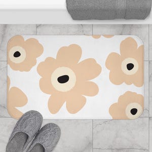 May include: A rectangular bath mat featuring a white background with a repeating pattern of large, stylised beige flowers. Each flower has rounded petals and a solid black circular centre. The mat has rounded corners and a soft, plush texture.