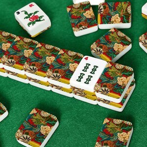 Puede incluir: Un conjunto de fichas de mahjong rectangulares con base blanca y marcas florales y de caracteres en verde y rojo, junto a fichas con un patr&oacute;n detallado y colorido de dragones y flores en tonos rojo, verde, amarillo y blanco.