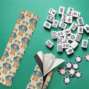 May include: A beige Mahjong tile case with a palm tree pattern, open to reveal a zipper. Scattered white Mahjong tiles with black and red markings, along with blue, red, and white game chips, are on a green surface.