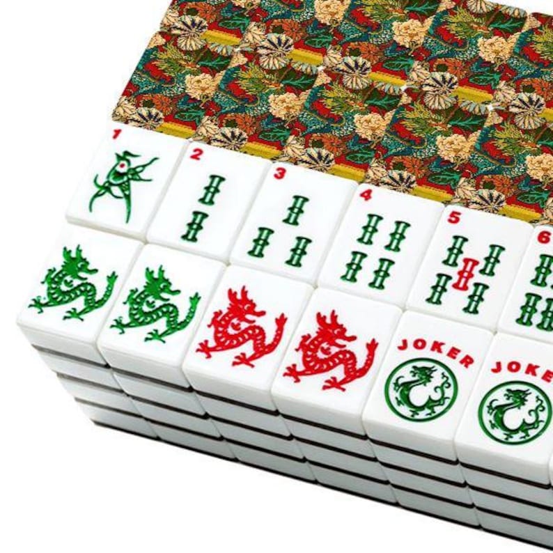 Puede incluir: Un conjunto de fichas de mahjong blancas dispuestas en filas. Las fichas presentan s&iacute;mbolos de drag&oacute;n grabados en verde y rojo, patrones de bamb&uacute; y el texto Joker con iconos de drag&oacute;n circulares. El reverso muestra un colorido patr&oacute;n floral.