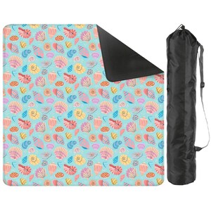 May include: A beach blanket featuring a seashell design in pink, yellow, and blue on a light blue backdrop. It has a black underside and is accompanied by a black carry bag with a shoulder strap.
