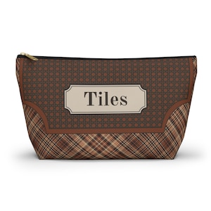 May include: A brown zippered pouch featuring a dark brown woven cane pattern on the upper section and a brown plaid pattern on the lower section. A central cream-colored rectangular label displays the word Tiles in dark brown lettering.