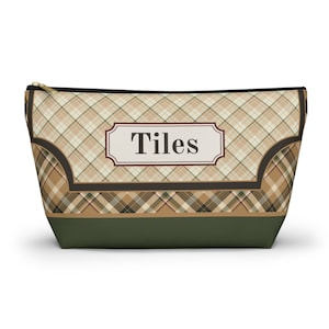 Puede incluir: Un estuche trapezoidal con cremallera que presenta un patrón de cuadros en beige, marrón y verde oscuro. Una etiqueta rectangular central con borde marrón oscuro contiene la palabra "Tiles" en una fuente serif oscura. La base es verde oscuro.